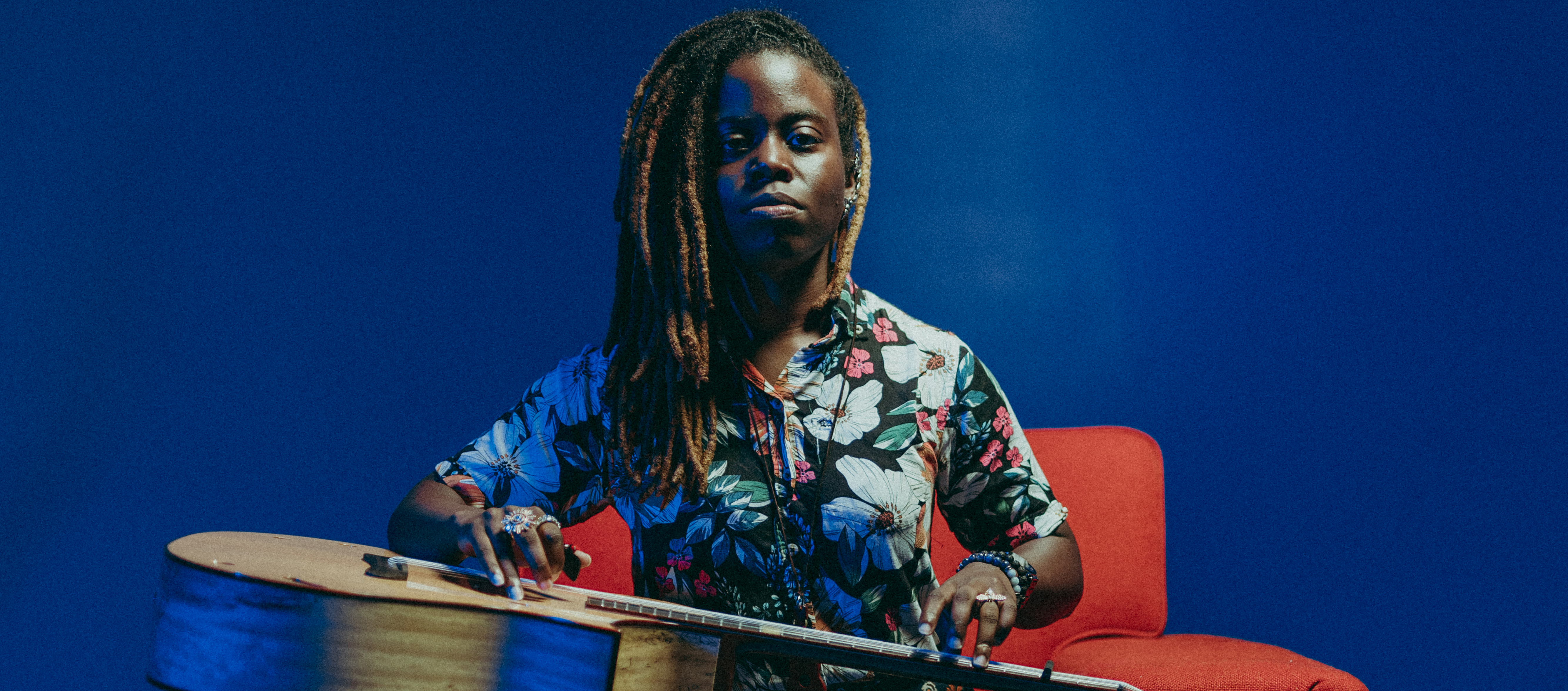 Musician with brown skin and ombre locs wears a floral-patterned outfit and sits on a red chair against a blue background with an acoustic guitar on her lap.