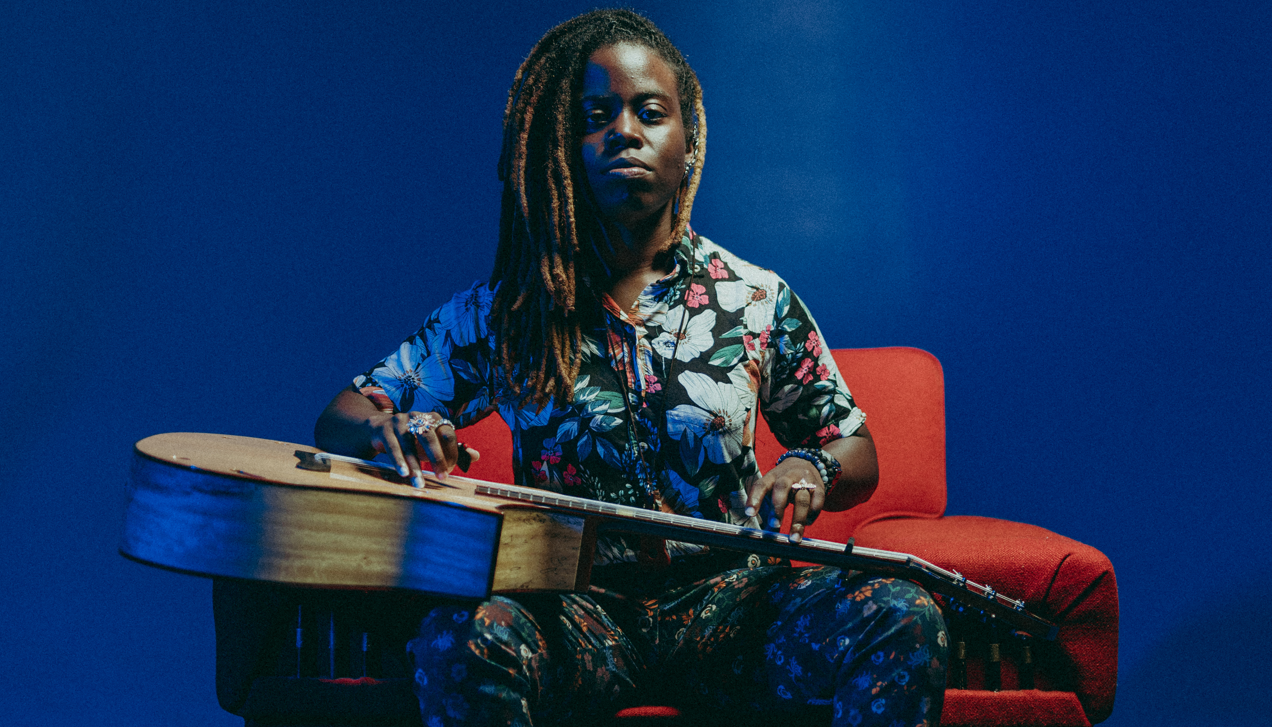 Musician with brown skin and ombre locs wears a floral-patterned outfit and sits on a red chair against a blue background with an acoustic guitar on her lap.
