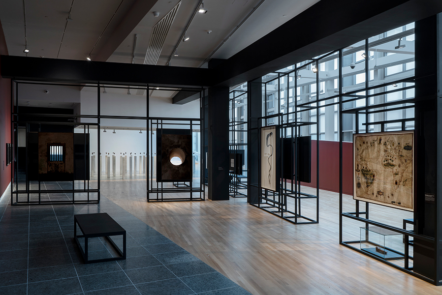 Darkened gallery space with black scaffolding structure that displays framed artworks. Light enters from a gridded glass wall in the background. 
