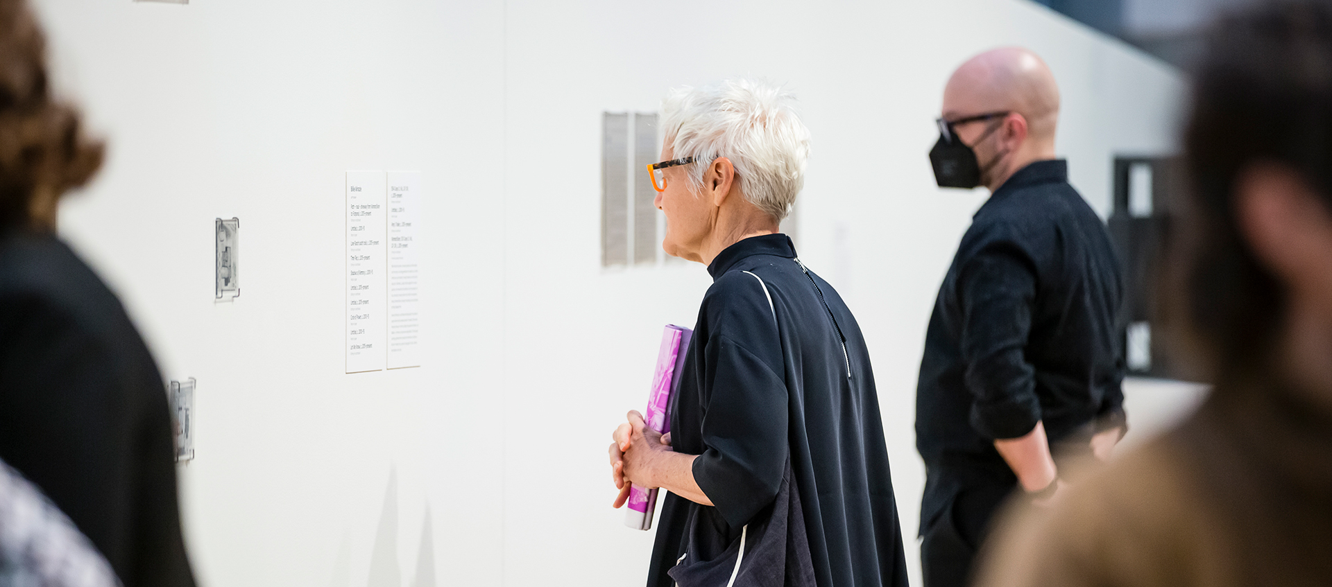 Two people read labels on a white gallery wall. The person in the foreground holds a rolled-up publication from the exhibition Sharing Circles: Carol Newhouse and the WomanShare Collective.
