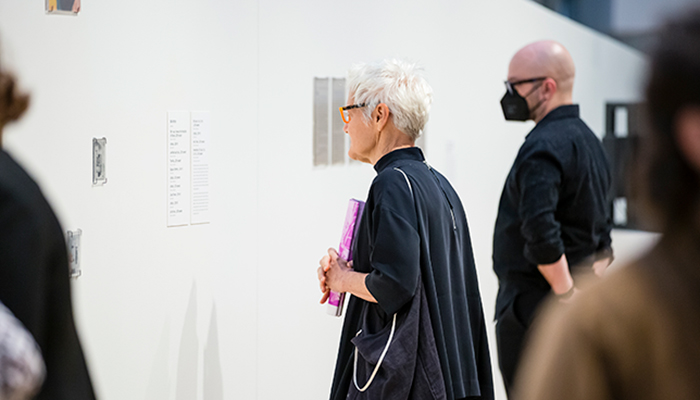 Two people read labels on a white gallery wall. The person in the foreground holds a rolled-up publication from the exhibition Sharing Circles: Carol Newhouse and the WomanShare Collective.