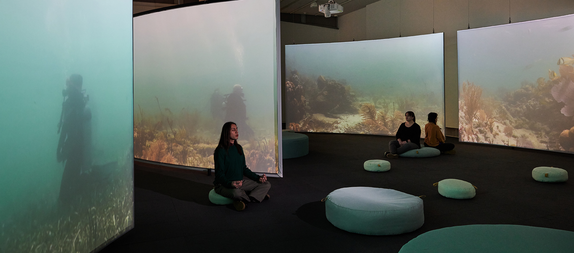 A dimly lit gallery space with four large screens projecting scuba divers meditating among coral reefs. Three visitors sit in meditation on round cushions on the floor.