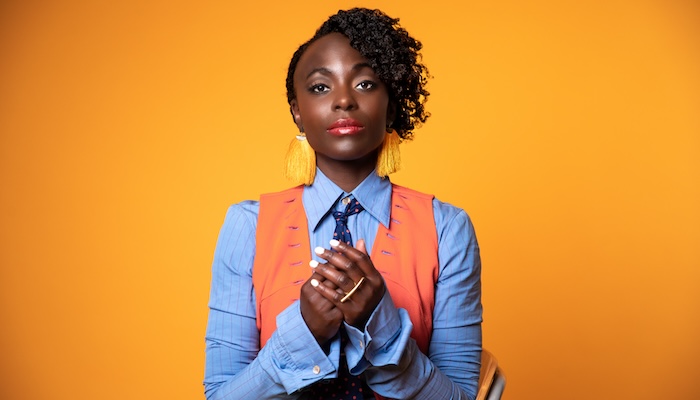 A woman with dark skin and short hair stands in front of an orange background with her hands clasped together as she stares at the camera. 