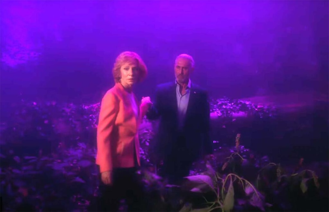 A man and a woman hold hands in a landscape lit in deep purple. The woman looks forward as the man looks at her.